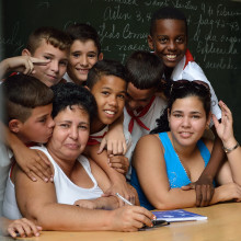 A teacher being hugged and kissed by her students while sitting on her desk.