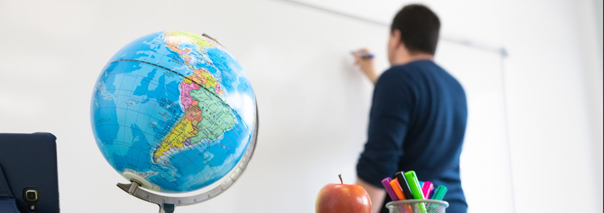 Teacher writing on a whiteboard. A globe and an apple over the desk. 